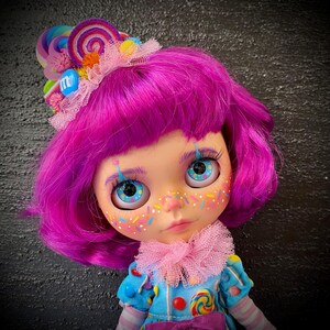 May include: A doll with bright purple hair and blue eyes. The doll has a whimsical face with colorful freckles and painted eyelashes. It wears a blue outfit with candy-themed embellishments and a pink ruffled collar. A candy-themed headpiece completes the look.