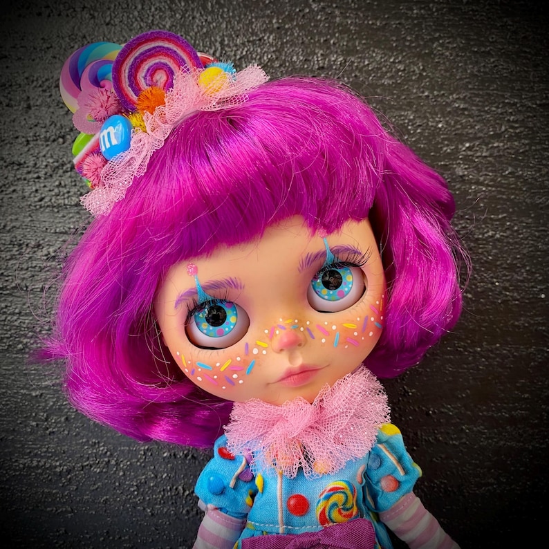 May include: A doll with bright purple hair and blue eyes. The doll has a whimsical face with colorful freckles and painted eyelashes. It wears a blue outfit with candy-themed embellishments and a pink ruffled collar. A candy-themed headpiece completes the look.