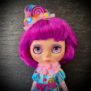 May include: A doll with bright pink hair and large eyes, wearing a blue dress with candy and lollipop details. The doll has a pink ruffled collar and a candy-themed headpiece with a variety of colorful candies.