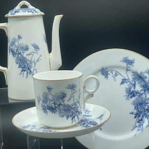 Peut inclure: Service à thé en porcelaine blanche avec motifs floraux bleus et bordures dorées. L'ensemble comprend une cafetière, une tasse, une soucoupe et une assiette. Le motif floral présente des fleurs et des feuilles bleues détaillées. L'ensemble est présenté sur une surface acrylique transparente.