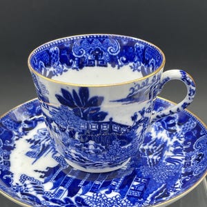Peut inclure: Un ensemble tasse et soucoupe bleu et blanc. La tasse et la soucoupe présentent un motif paysager détaillé avec un bord doré. Le motif est un motif bleu et blanc classique, parfait pour le thé ou le café.
