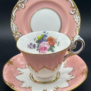 Könnte beinhalten: Ein rosafarbenes Tee-Set mit Blumendekor und goldenen Verzierungen. Die Teetasse hat ein zartes Blumenmuster mit rosa, orangefarbenen und gelben Blüten. Die Untertasse hat einen gewellten Rand und einen goldenen Rand.