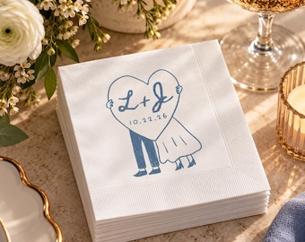 May include: A stack of white paper napkins featuring a blue ink illustration of two figures holding a large heart. The heart contains the letters L + J and the date 10.22.26. The napkins have a textured border and a smooth center panel.