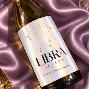 以下が含まれることがあります： 金色のてんびん座の星座グラフィック、LIBRA SEASONという文字、そしてHAPPY BIRTHDAY, LAUREN!というパーソナライズされたメッセージが書かれたホログラムラベル付きのワインボトル。ラベルは反射する虹色の仕上げになっています。