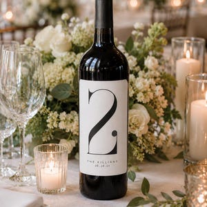 May include: A dark glass wine bottle featuring a white rectangular label with a large black number 2, the text THE KILLIANS, and the date 06.26.27. The bottle has a black cap and is designed as a table number for an event or wedding reception.