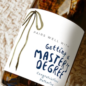 May include: A glass bottle with a white label featuring a watercolor olive green ribbon illustration. The text reads: PAIRS WELL WITH Getting a MASTER'S DEGREE Congratulations Katherine. The bottle contains a dark amber liquid.