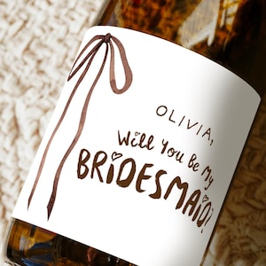 Puede incluir: Una botella de vidrio marrón con una etiqueta blanca. La etiqueta presenta el texto "OLIVIA, Will You Be My BRIDESMAID?" en escritura marrón, junto con un diseño de lazo marrón. La botella está sobre una superficie texturizada.