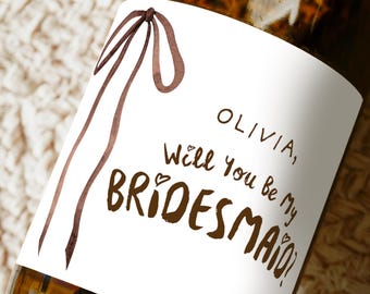 May include: A brown glass bottle with a white label. The label features the text "OLIVIA, Will You Be My BRIDESMAID?" in brown script, along with a brown ribbon design. The bottle is on a textured surface.