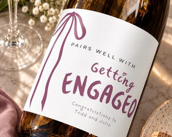 May include: A brown glass wine bottle with a white label featuring a purple ribbon illustration and the text: PAIRS WELL WITH Getting ENGAGED Congratulations to Todd and Julia. The label is printed in a mix of serif and script fonts.