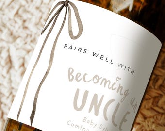 Può includere: Una bottiglia di vetro con un'etichetta bianca che presenta un'illustrazione di un nastro ad acquerello marrone e testo nero che recita: PAIRS WELL WITH becoming an UNCLE Baby Sylva Coming June 2026. L'etichetta include il contorno di un calice.