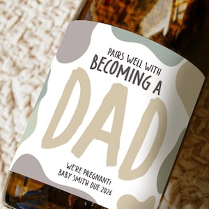 May include: A bottle with a custom label reading "PAIRS WELL WITH BECOMING A DAD." The label also includes the text "WE'RE PREGNANT! BABY SMITH DUE 2026" in a playful font. The label has a white background with abstract shapes in muted colours.