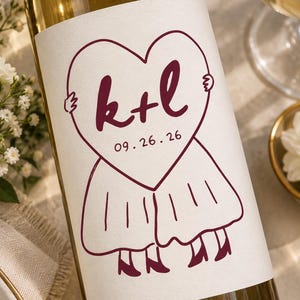 May include: A clear glass wine bottle with a white rectangular label featuring a dark red line drawing of a heart held by two figures wearing skirts and heels. The text k+l and the date 09.26.26 are printed in dark red inside the heart shape.