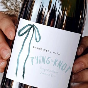 May include: A wine bottle with a white rectangular label featuring a watercolour-style teal ribbon illustration. The text reads PAIRS WELL WITH TYING the KNOT, followed by Congratulations Jemma & Eric in a light blue script font.