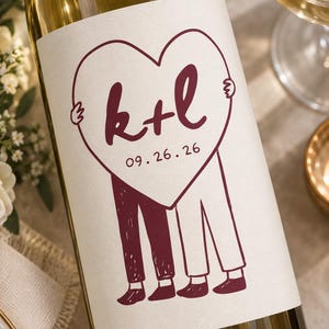 May include: A glass wine bottle with a white label featuring a dark red line drawing of two figures holding a large heart. The heart contains the letters k+l and the date 09.26.26. The figures are depicted from the waist down, wearing trousers and shoes.