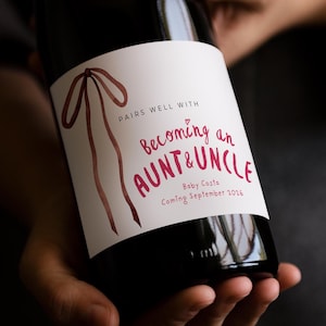 Puede incluir: Una botella de vino de vidrio oscuro con una etiqueta rectangular blanca que presenta una ilustraci&oacute;n de cinta en acuarela marr&oacute;n y texto rojo: PAIRS WELL WITH Becoming an AUNT & UNCLE Baby Costa Coming September 2026 con un peque&ntilde;o coraz&oacute;n rojo.