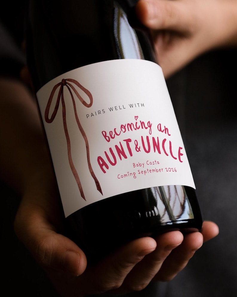 Puede incluir: Una botella de vino de vidrio oscuro con una etiqueta rectangular blanca que presenta una ilustraci&oacute;n de cinta en acuarela marr&oacute;n y texto rojo: PAIRS WELL WITH Becoming an AUNT & UNCLE Baby Costa Coming September 2026 con un peque&ntilde;o coraz&oacute;n rojo.