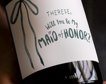 May include: A white wine bottle label featuring a dark green hand-drawn bow illustration on the left side. The text in dark green reads: Therese, Will You Be My MAID of HONOR? The label is applied to a dark glass bottle with a smooth finish.
