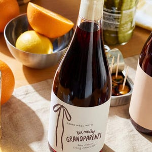 Puede incluir: Una botella de vidrio llena de l&iacute;quido rojo oscuro, sellada con un tap&oacute;n de corcho. La etiqueta blanca presenta un dibujo lineal marr&oacute;n de una cinta y el texto: PAIRS WELL WITH Becoming GRANDPARENTS Baby Johnson Coming January 2027.