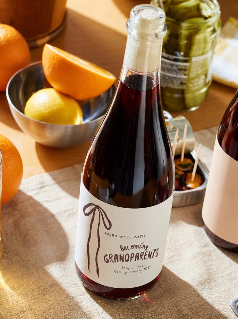 Puede incluir: Una botella de vidrio llena de l&iacute;quido rojo oscuro, sellada con un tap&oacute;n de corcho. La etiqueta blanca presenta un dibujo lineal marr&oacute;n de una cinta y el texto: PAIRS WELL WITH Becoming GRANDPARENTS Baby Johnson Coming January 2027.