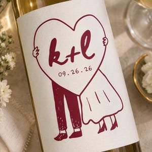 May include: A glass wine bottle with a white label featuring a dark red line drawing of two figures holding a large heart. The heart contains the letters k+l and the date 09.26.26. The figures are depicted in a simple, sketch-like style.