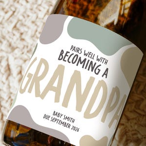 May include: A champagne bottle with a custom label. The label says "Pairs well with Becoming a Grandpa", with the baby's name and due date. The label has a neutral colour palette with abstract shapes.