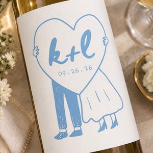 May include: A glass wine bottle featuring a white label with a light blue illustration of two figures holding a large heart. The heart contains the blue text k+l and the date 09.26.26. The figures are depicted from the waist down in blue.