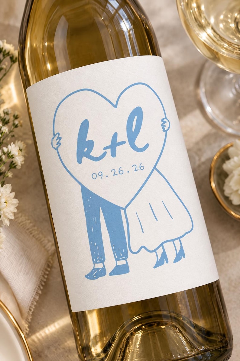 May include: A glass wine bottle featuring a white label with a light blue illustration of two figures holding a large heart. The heart contains the blue text k+l and the date 09.26.26. The figures are depicted from the waist down in blue.