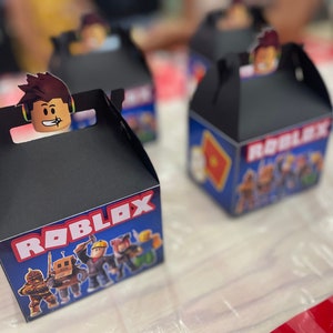 May include: Black favor boxes with a Roblox theme. The boxes feature a blue background with a red and white Roblox logo and images of Roblox characters. The boxes have a handle on the top and are perfect for holding small gifts or treats.