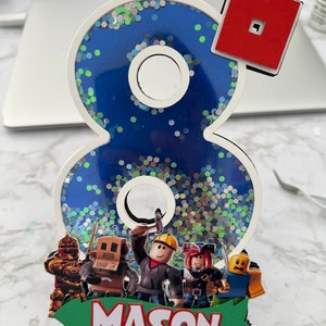 May include: A blue and white glittery number eight cake topper with a red Roblox logo, a green banner with the name "MASON" and four Roblox characters.