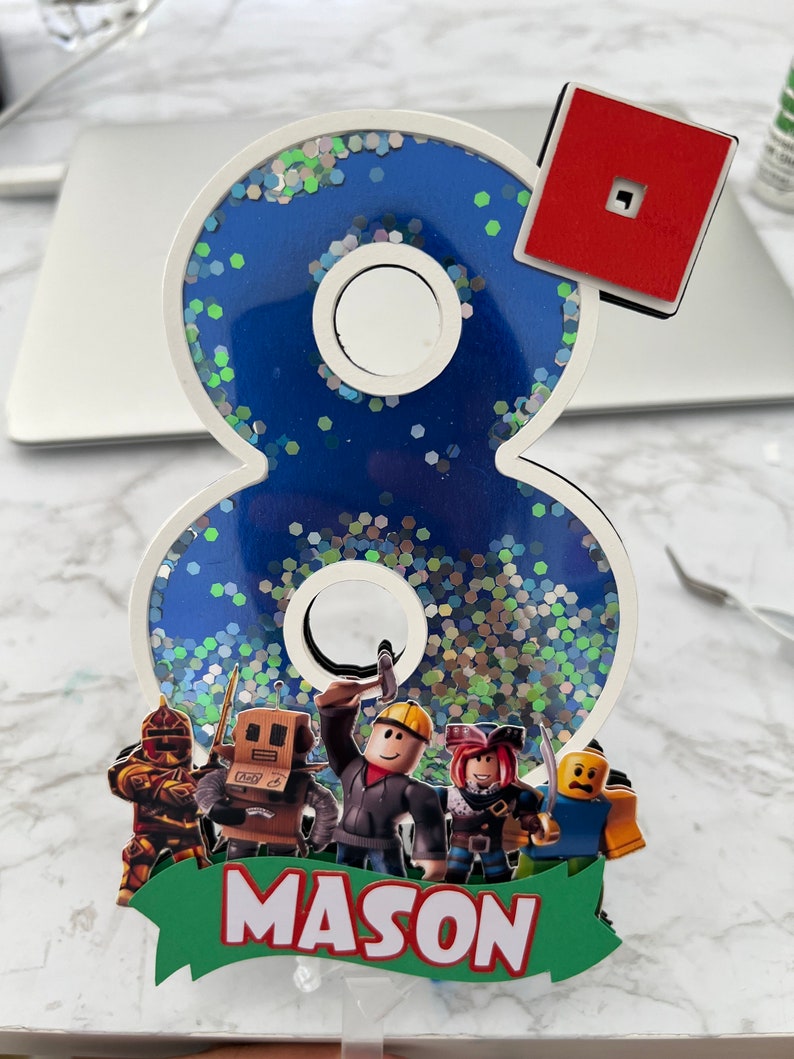 May include: A blue and white glittery number eight cake topper with a red Roblox logo, a green banner with the name "MASON" and four Roblox characters.