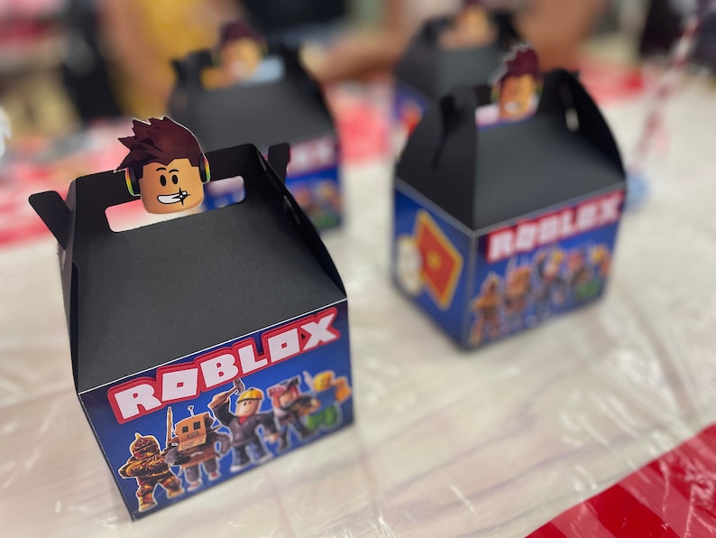 May include: Black favor boxes with a Roblox theme. The boxes feature a blue background with a red and white Roblox logo and images of Roblox characters. The boxes have a handle on the top and are perfect for holding small gifts or treats.