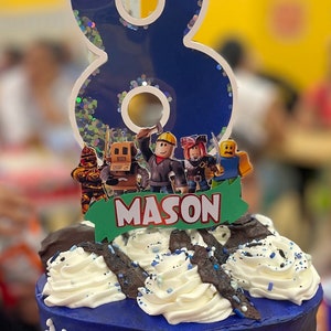 May include: A blue and white birthday cake with a blue number 8 candle and a Roblox themed topper. The cake is decorated with white frosting and blue sprinkles. The topper features a banner that says "MASON".