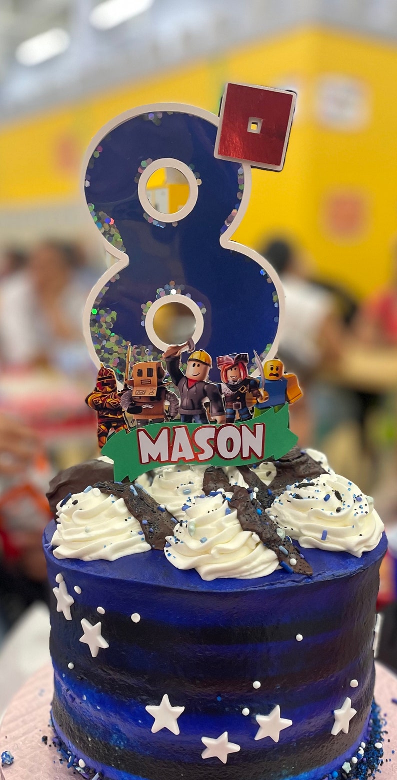May include: A blue and white birthday cake with a blue number 8 candle and a Roblox themed topper. The cake is decorated with white frosting and blue sprinkles. The topper features a banner that says "MASON".