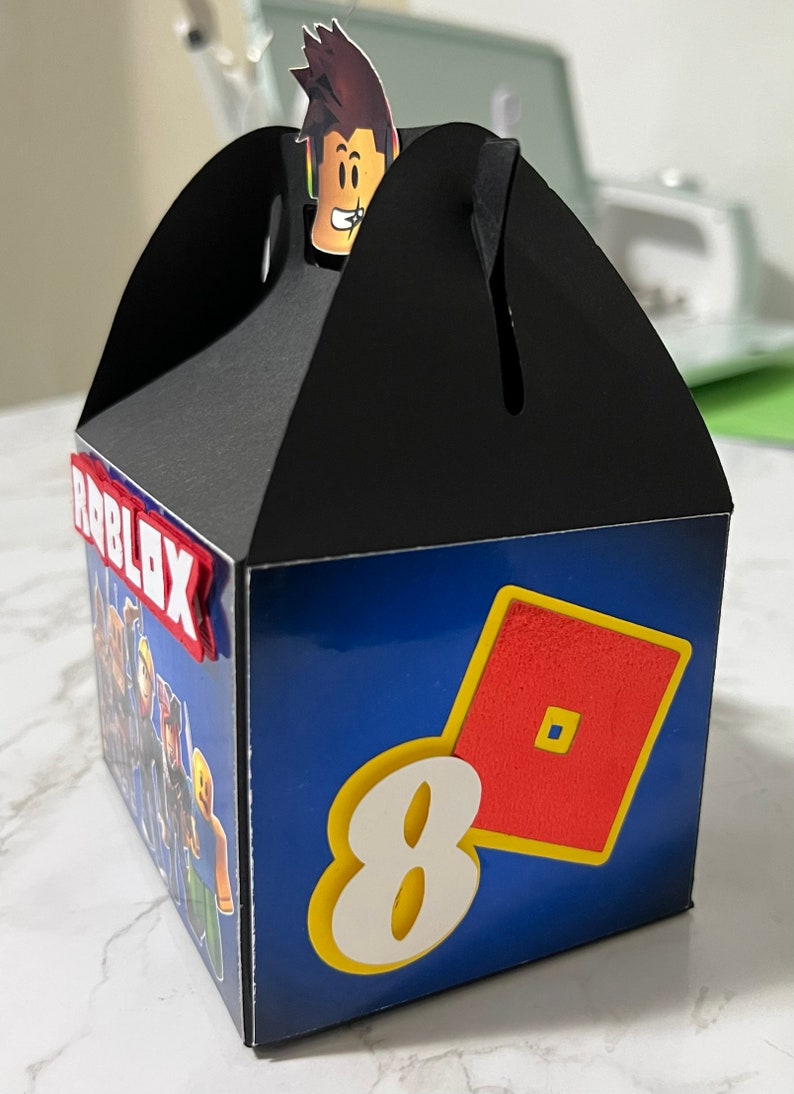 May include: A black and blue favor box with a Roblox logo, a red and yellow Roblox logo, and the number 8. The box has a black top with a cut-out for a handle. A small cartoon character with brown hair is on top of the box.