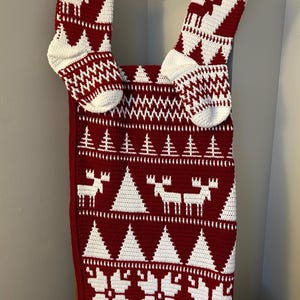 May include: Handmade Christmas stocking and matching banner in red and white crochet. The design features reindeer, trees, and geometric patterns. The stocking is hanging on a wooden ladder. A festive, cozy holiday decoration.