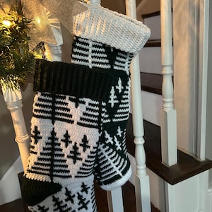 Pode incluir: Duas meias de Natal feitas à mão. As meias são brancas com um padrão de árvore verde e punhos verdes. As meias estão penduradas em um corrimão branco de escada. Uma decoração festiva.