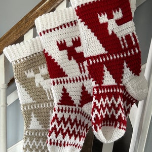 Peut inclure: Trois bas de Noël faits à la main, crochetés avec du fil blanc, rouge et beige. Chaque bas présente un motif festif différent, notamment un élan, des arbres de Noël et des motifs géométriques. Les bas sont accrochés sur un escalier en bois.