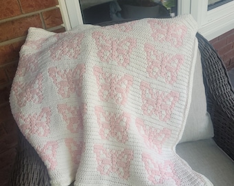 May include: A white crocheted blanket with a pink butterfly pattern. The blanket is folded and placed on a wicker chair.