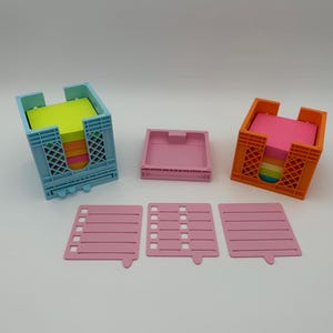 May include: Two colorful plastic note holder organizers with removable trays. The organizers are in the shape of a crate and are filled with colorful sticky notes. The organizers are on a white surface with three pink notepads.