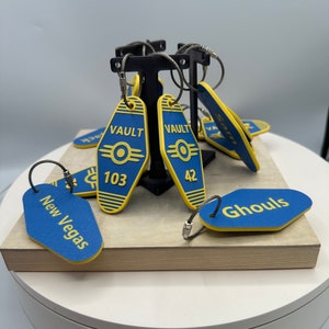 May include: A set of eight Fallout themed keychains. The keychains are shaped like the Vault-Tec door signs and feature different Vault numbers, including Vault 103, Vault 42, and "Ghouls". The keychains are yellow and blue with black text and a silver chain.