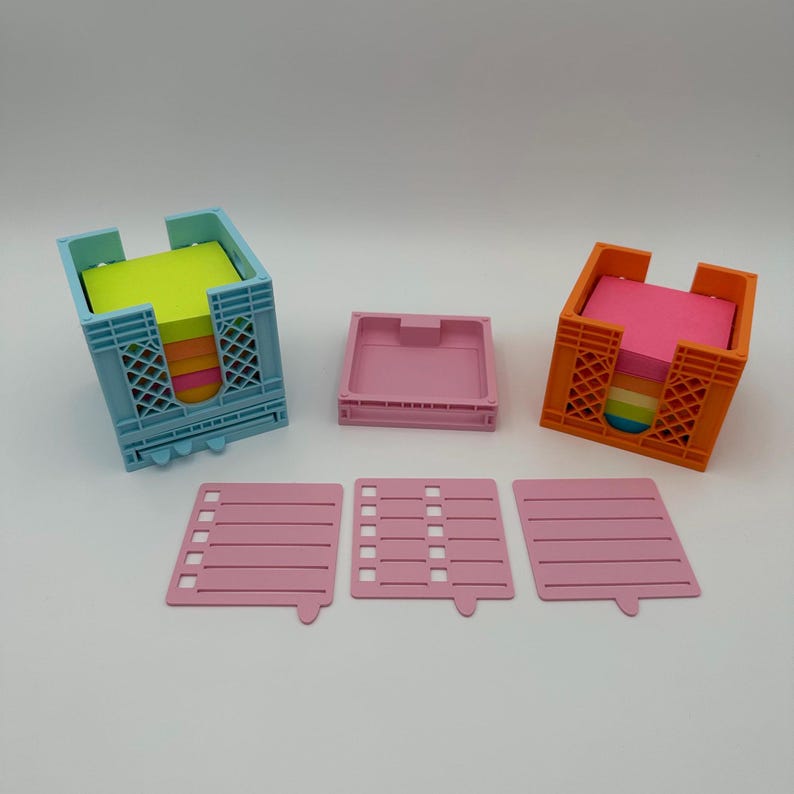 May include: Two colorful plastic note holder organizers with removable trays. The organizers are in the shape of a crate and are filled with colorful sticky notes. The organizers are on a white surface with three pink notepads.