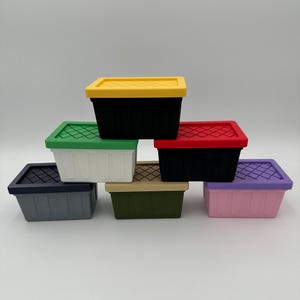 May include: A collection of small, colorful storage bins. The bins are rectangular with a textured lid and come in various colors, including black, white, green, yellow, red, pink, and olive green. The lids have a grid-like pattern.