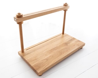 May include: A wooden book press with a long, narrow base and a screw mechanism for clamping books. The press is made of light-colored wood and has a simple, functional design.