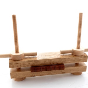 May include: A wooden book press with two wooden screws and a book in the center. The press is made of light brown wood and has a simple design.