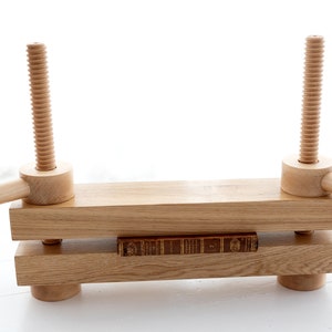 May include: A wooden book press with two adjustable screws and handles. A book is being pressed between the two wooden beams.