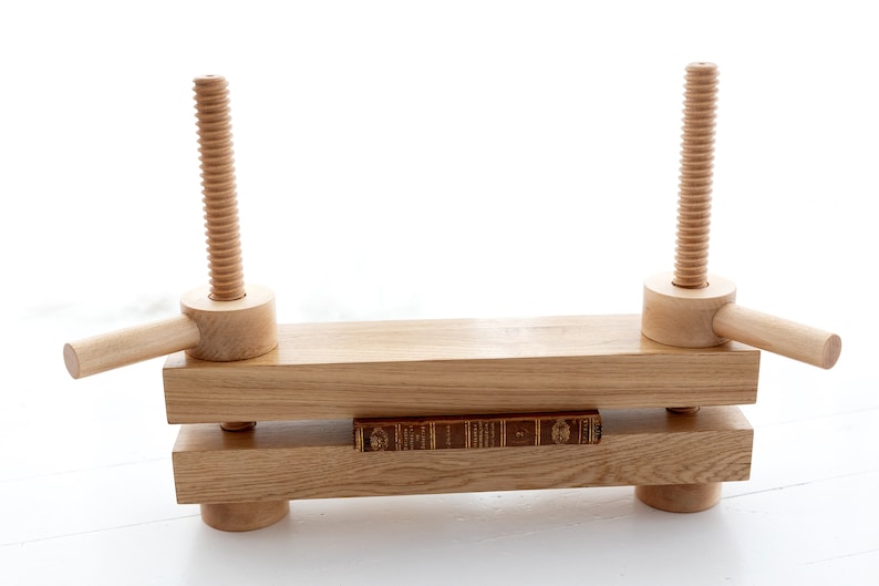 May include: A wooden book press with two adjustable screws and handles. A book is being pressed between the two wooden beams.