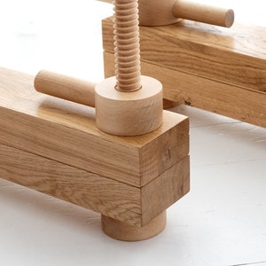 May include: A wooden clamp with a threaded screw and two handles. The clamp is made of natural wood and has a rustic look.