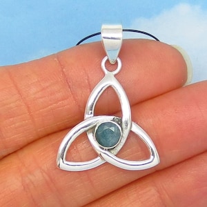 May include: A silver-colored pendant with a Celtic knot design. The pendant features a central, round, teal-colored stone. The pendant is suspended from a black cord, and is held in a hand against a light blue background.