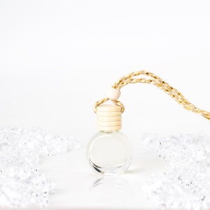 Könnte beinhalten: Kleine, klare Glasflasche mit Holzverschluss und geflochtener gold-beiger Kordel zum Aufhängen. Die Flasche ist rund und leer, mit einem hellen Holzverschluss. Die Kordel ist mit einer kleinen Holzperle am Verschluss befestigt.