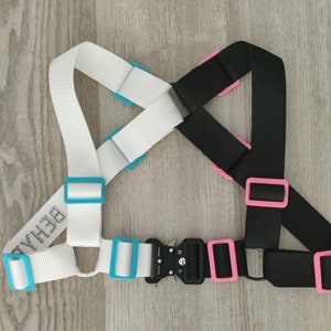 May include: A white and black adjustable harness with blue and pink buckles. The white strap has the word "BEHAVE" printed on it.